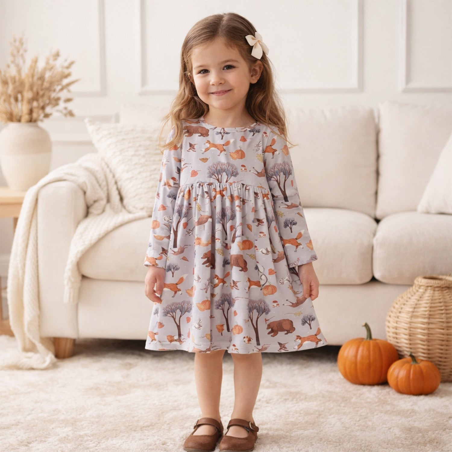 A young girl stands in a cozy living room wearing a long-sleeve dress with an all-over woodland animal print featuring foxes, deer, and trees on a light gray background. She is wearing brown mule shoes, with a cream couch, pumpkins, and a wicker basket in the background.