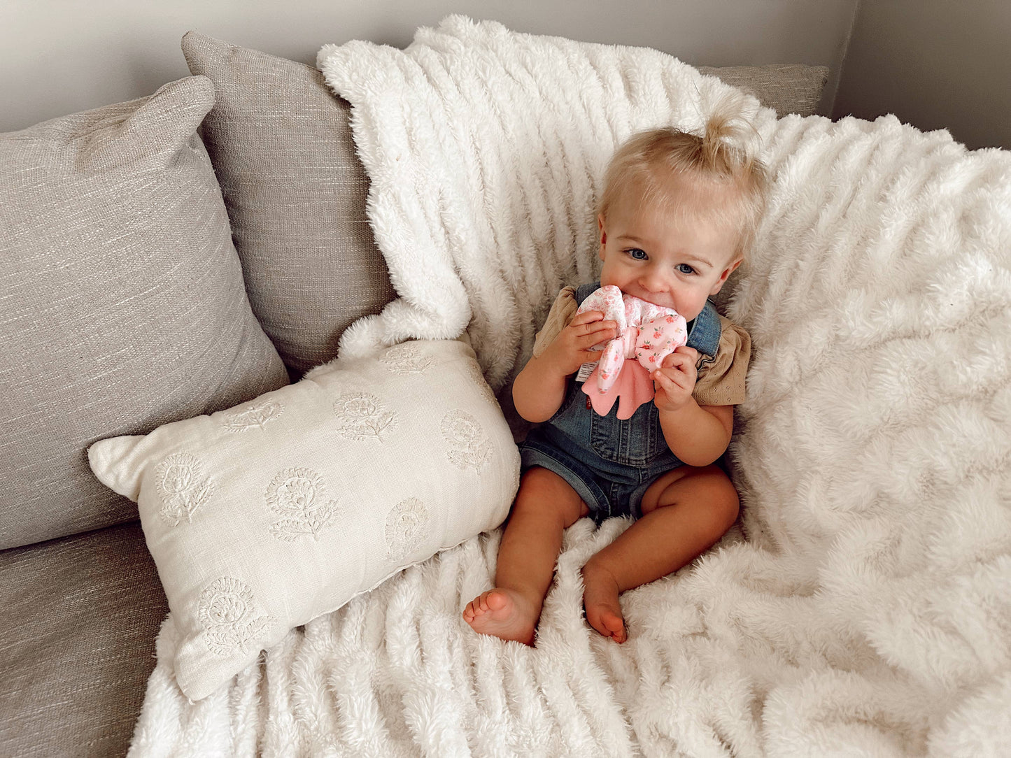 Child sitting on a couch with a white blanket and pillows, holding a pink toy.