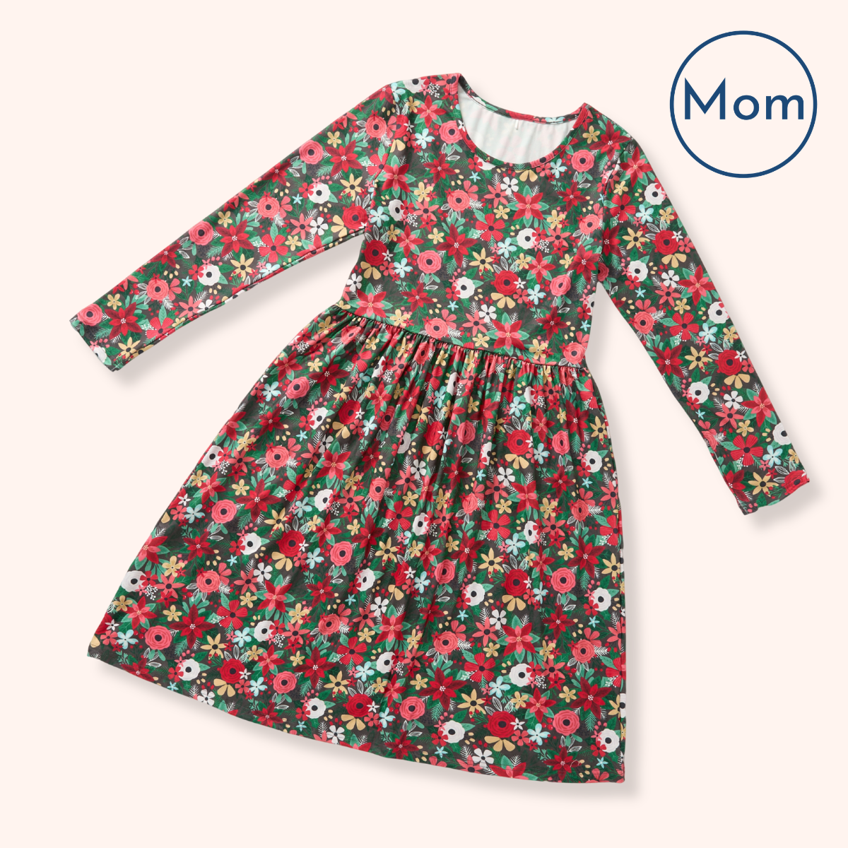 A long-sleeve girls' dress with a dense floral print in red, pink, white, and yellow on a dark green background, shown on a light beige background with a 'Mom' badge in the top right corner.