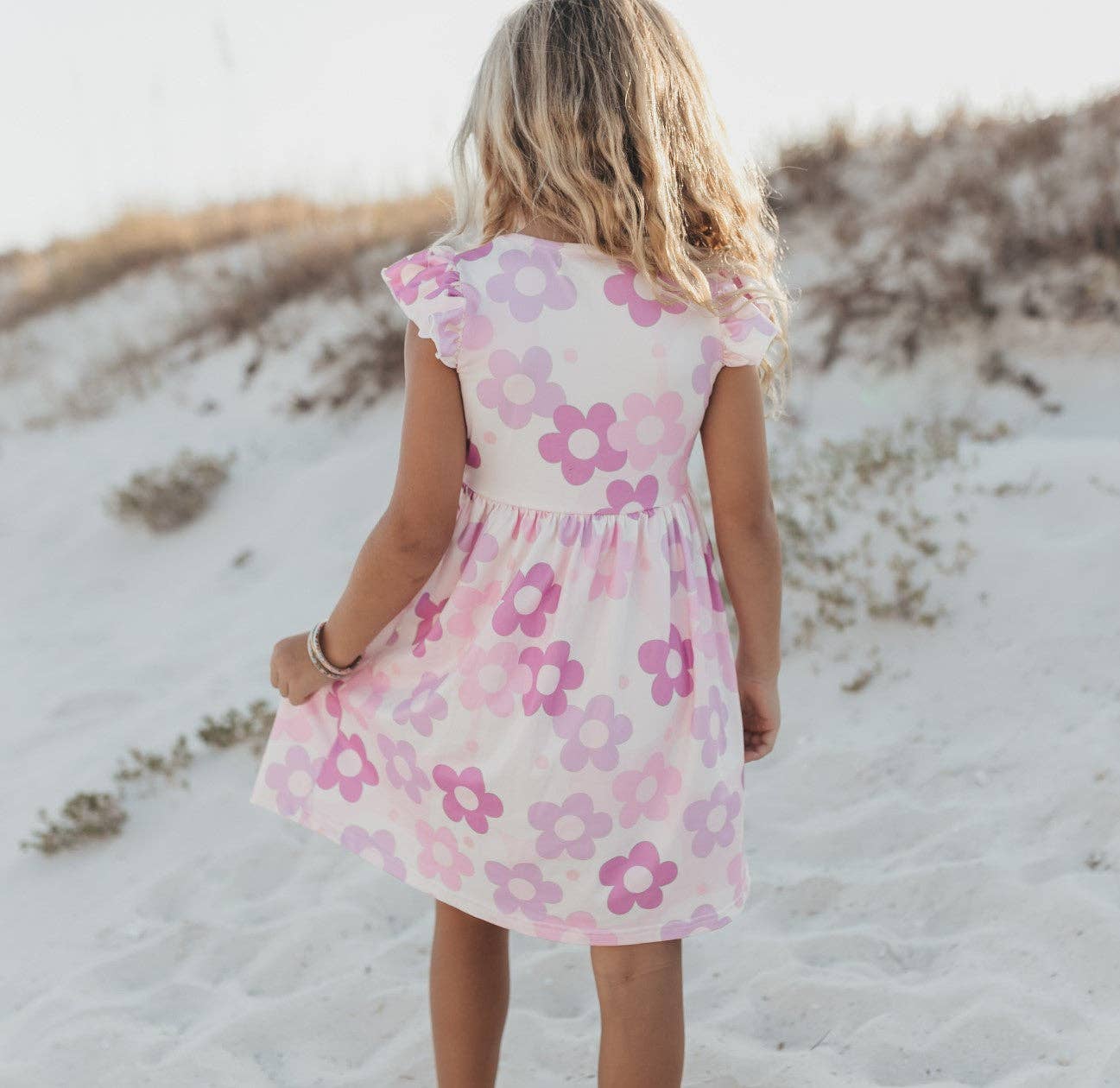 Child wearing a pink floral dress standing on a sandy beach.