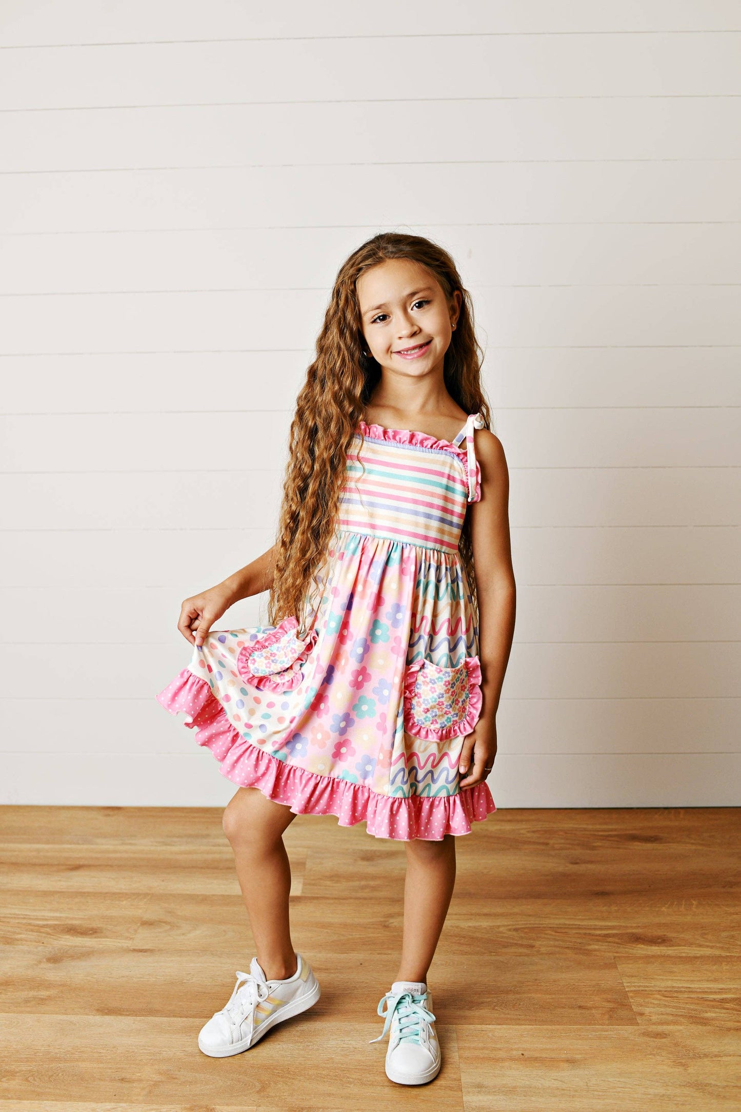 Young girl wearing a colorful dress standing on a wooden floor with a white wall background