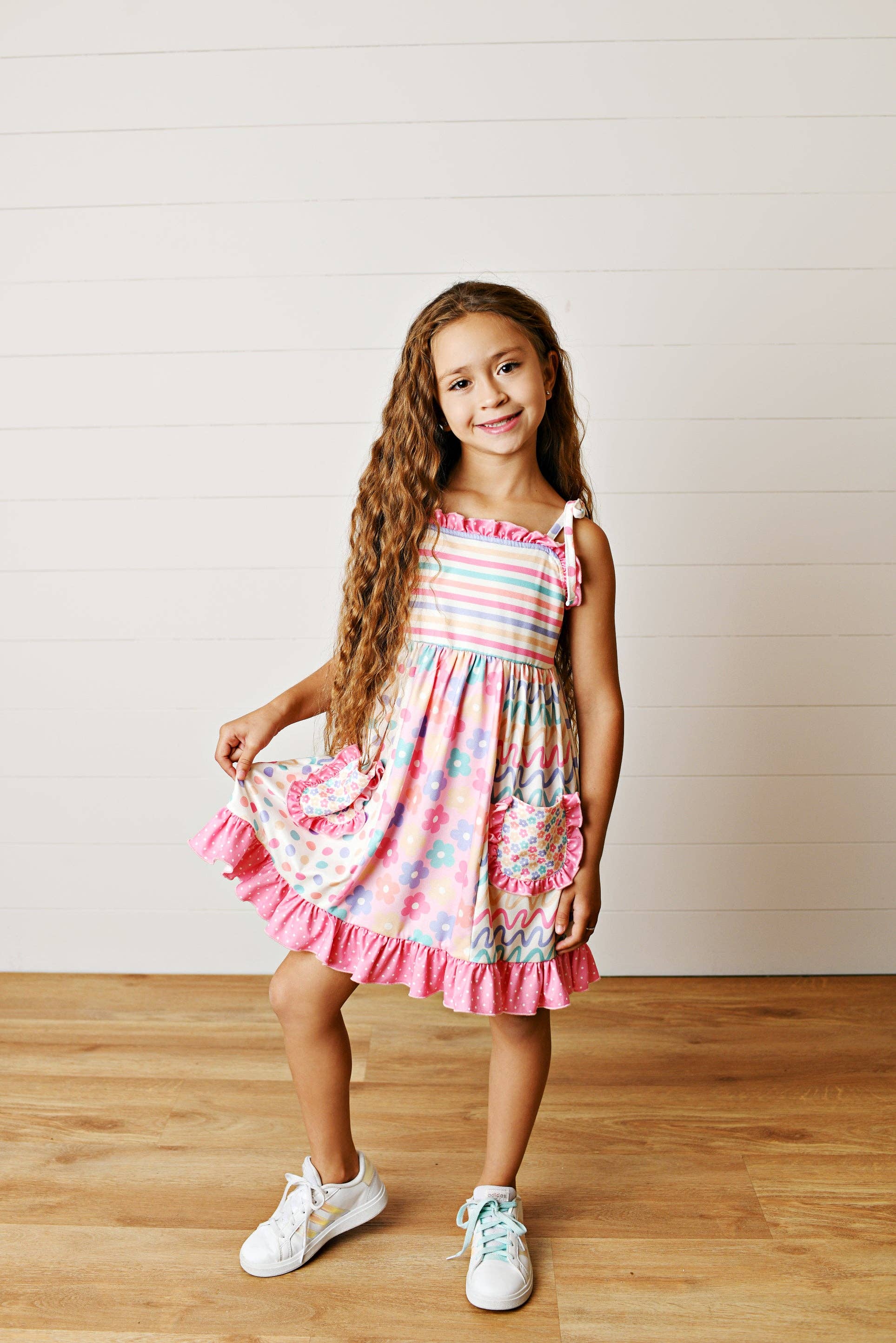 Young girl wearing a colorful dress standing on a wooden floor with a white wall background