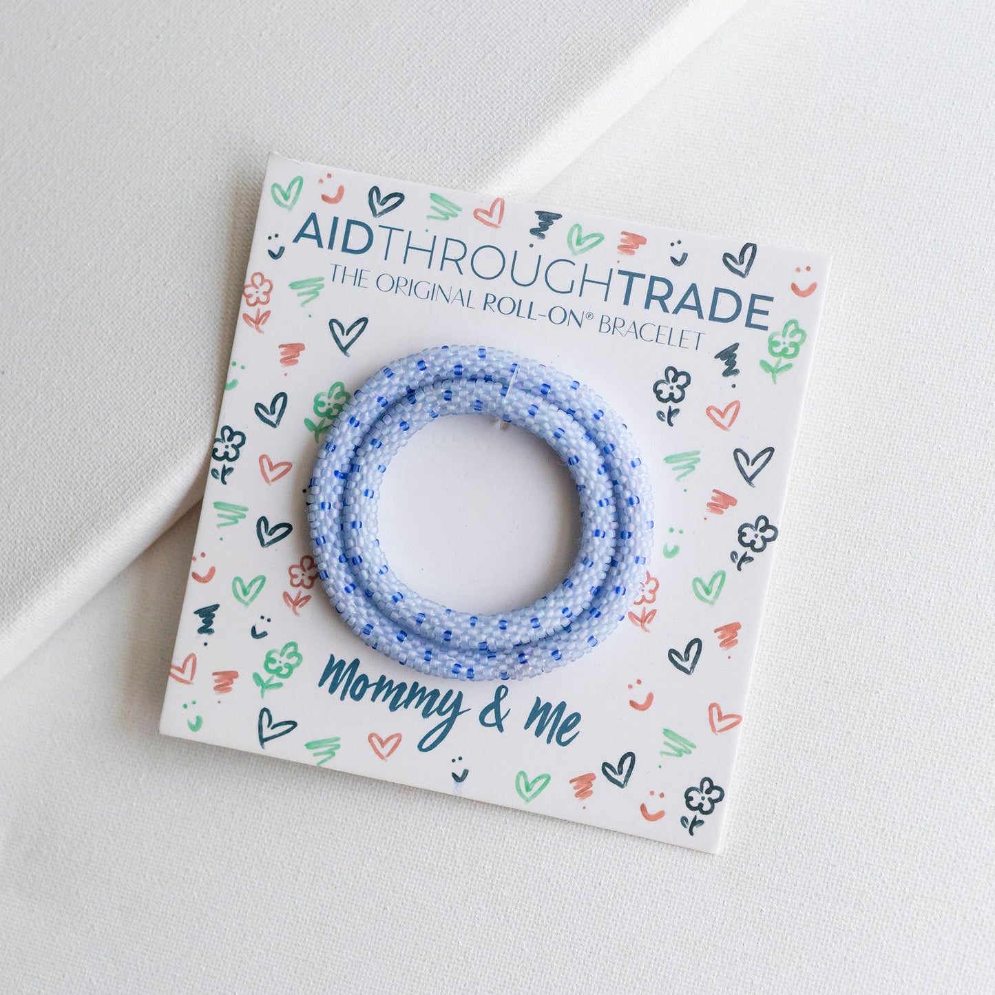Blue bracelet on a colorful 'Aid Through Trade' card with 'Mommy & Me' text on a light gray background