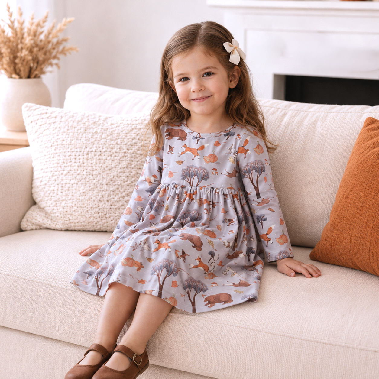 A young girl with a white bow in her hair sits on a beige sofa wearing a long-sleeve dress with an autumn woodland print of foxes, deer, and trees. The dress is a dusty lavender-gray color, and the background features a neutral living room setting with a white fireplace and decorative pillows.
