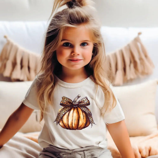 Child wearing a white t-shirt with a pumpkin design, sitting on a couch.