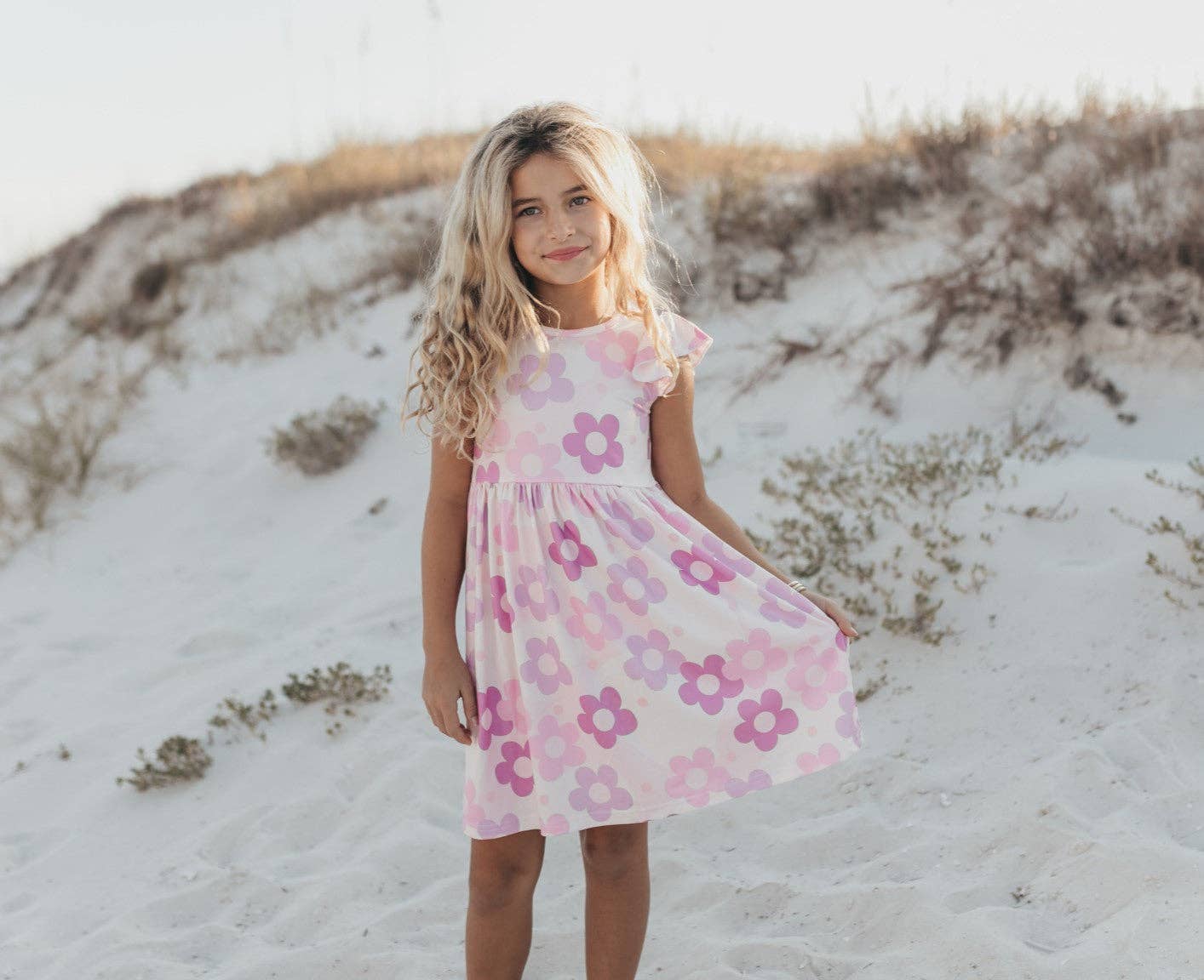 Young girl in a pink floral dress standing on a sandy beach.