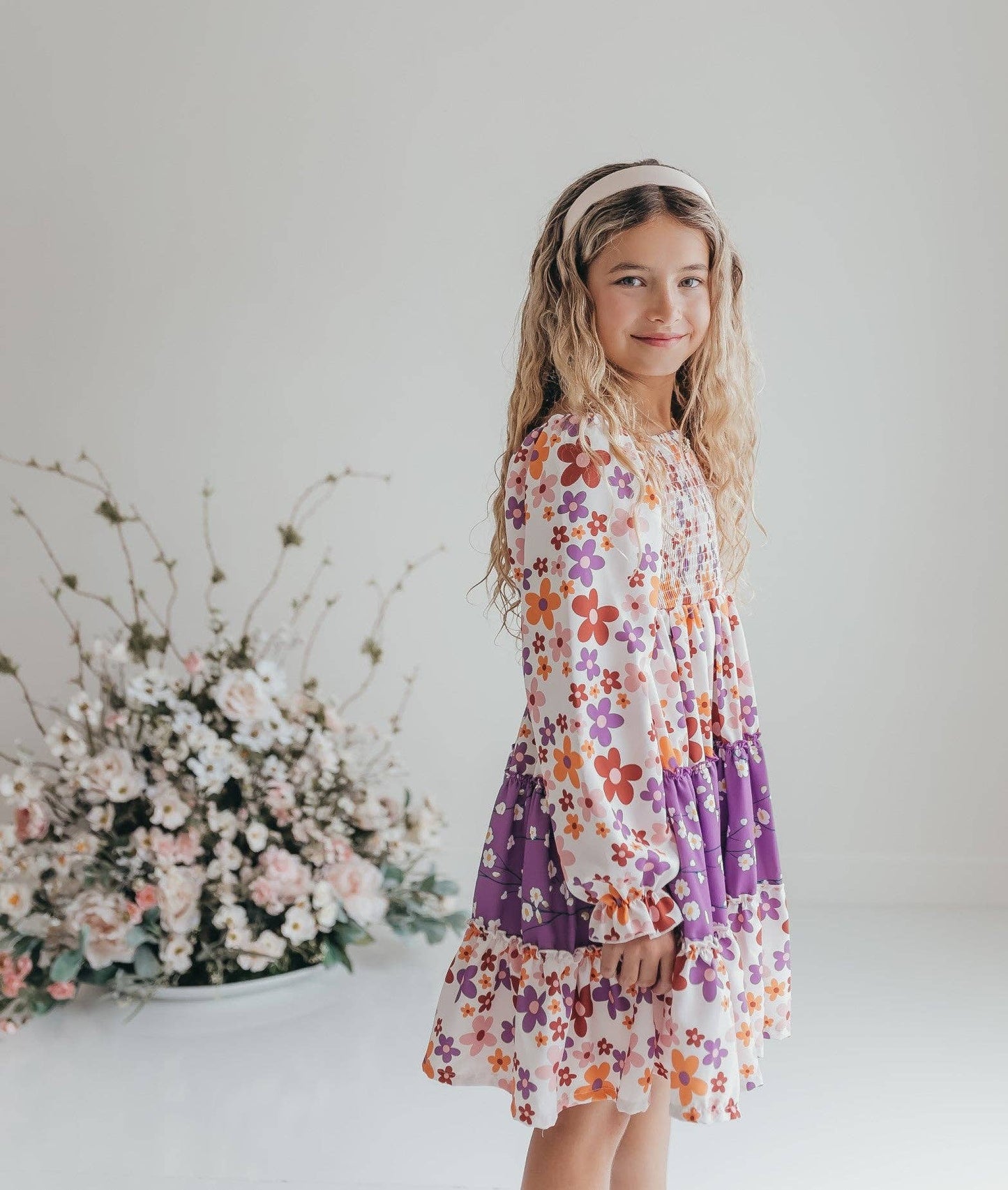 Young girl in a floral dress standing next to flowers against a plain background