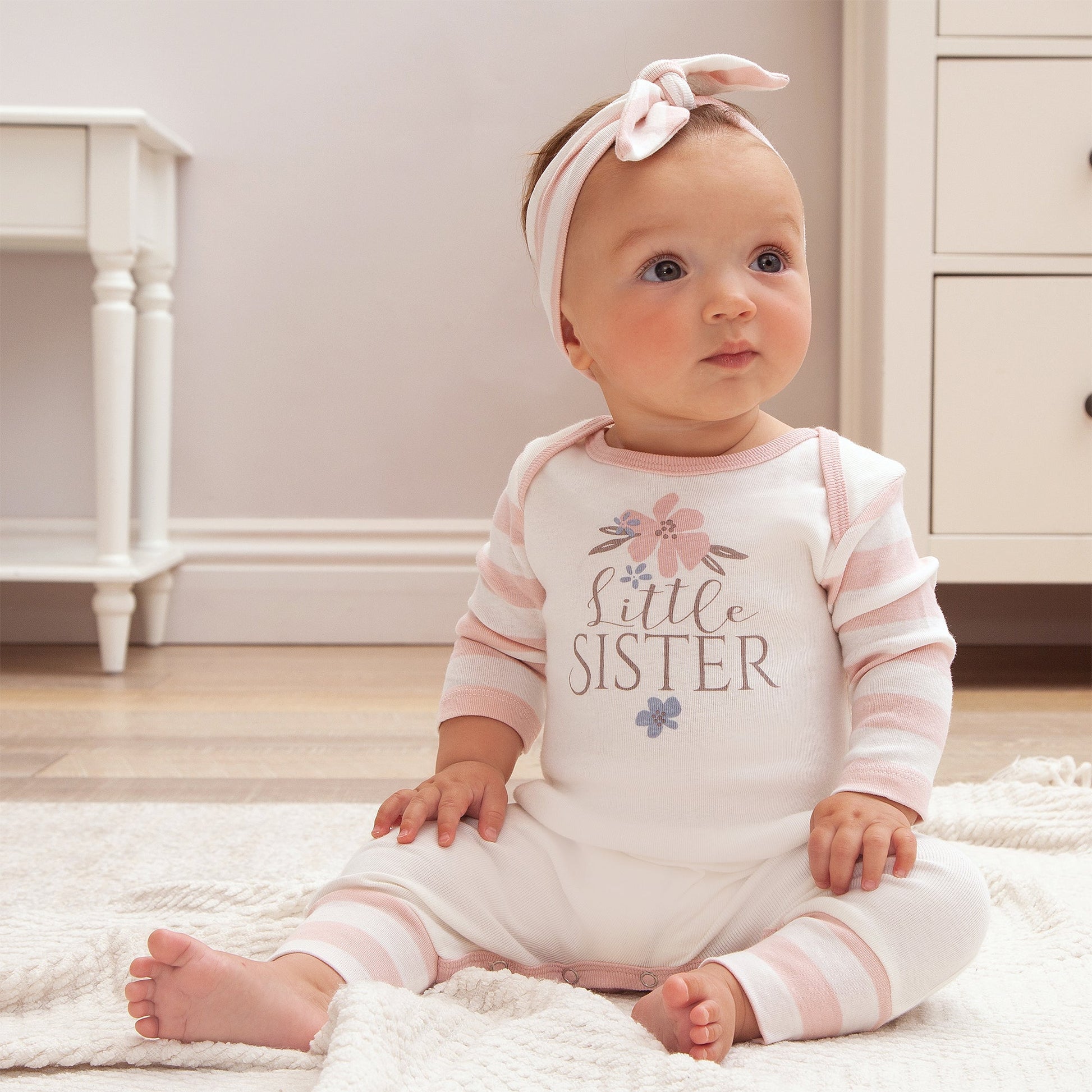 Baby wearing a 'Little Sister' outfit sitting on a rug.