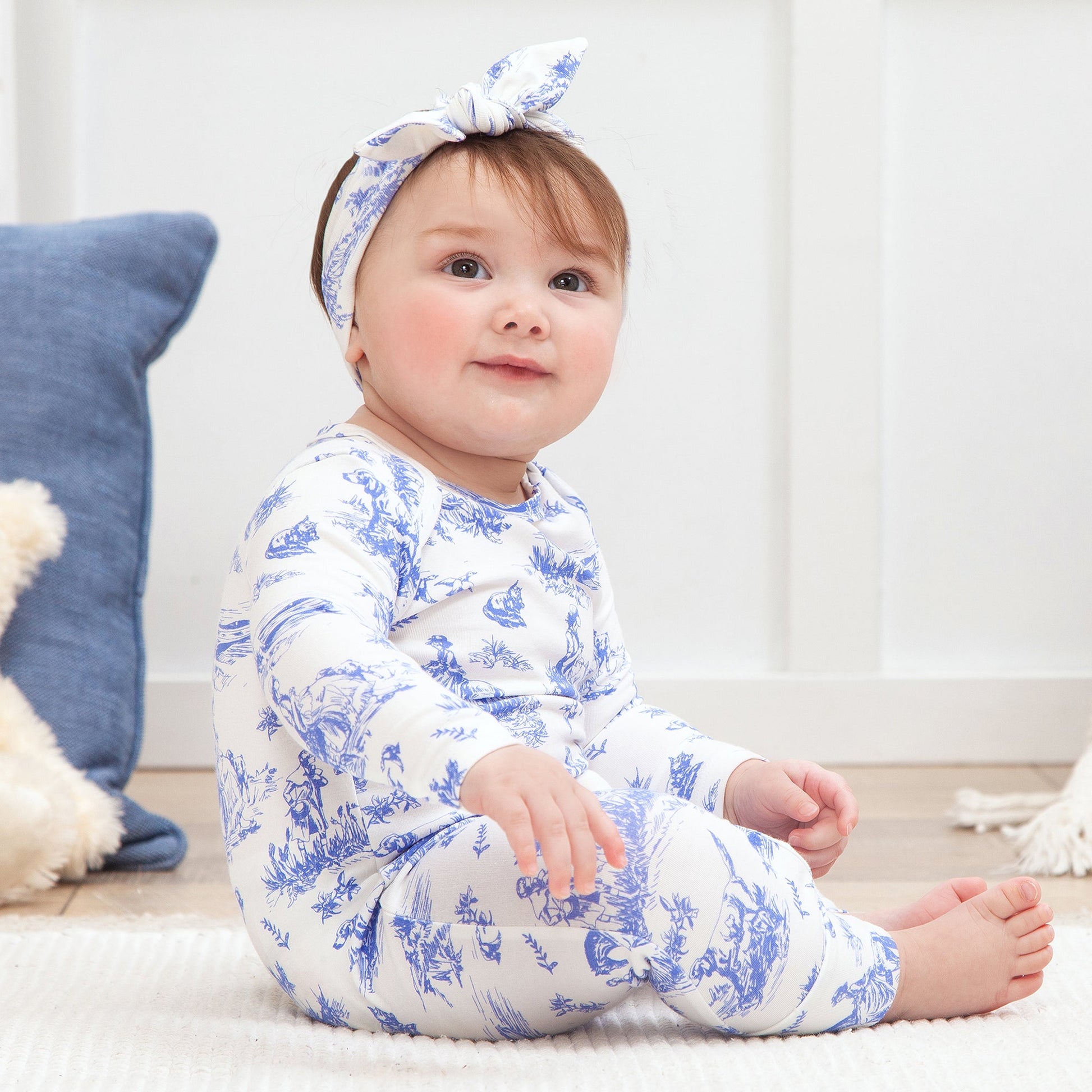 Baby wearing a blue and white patterned outfit with a matching headband, sitting on a wooden floor.