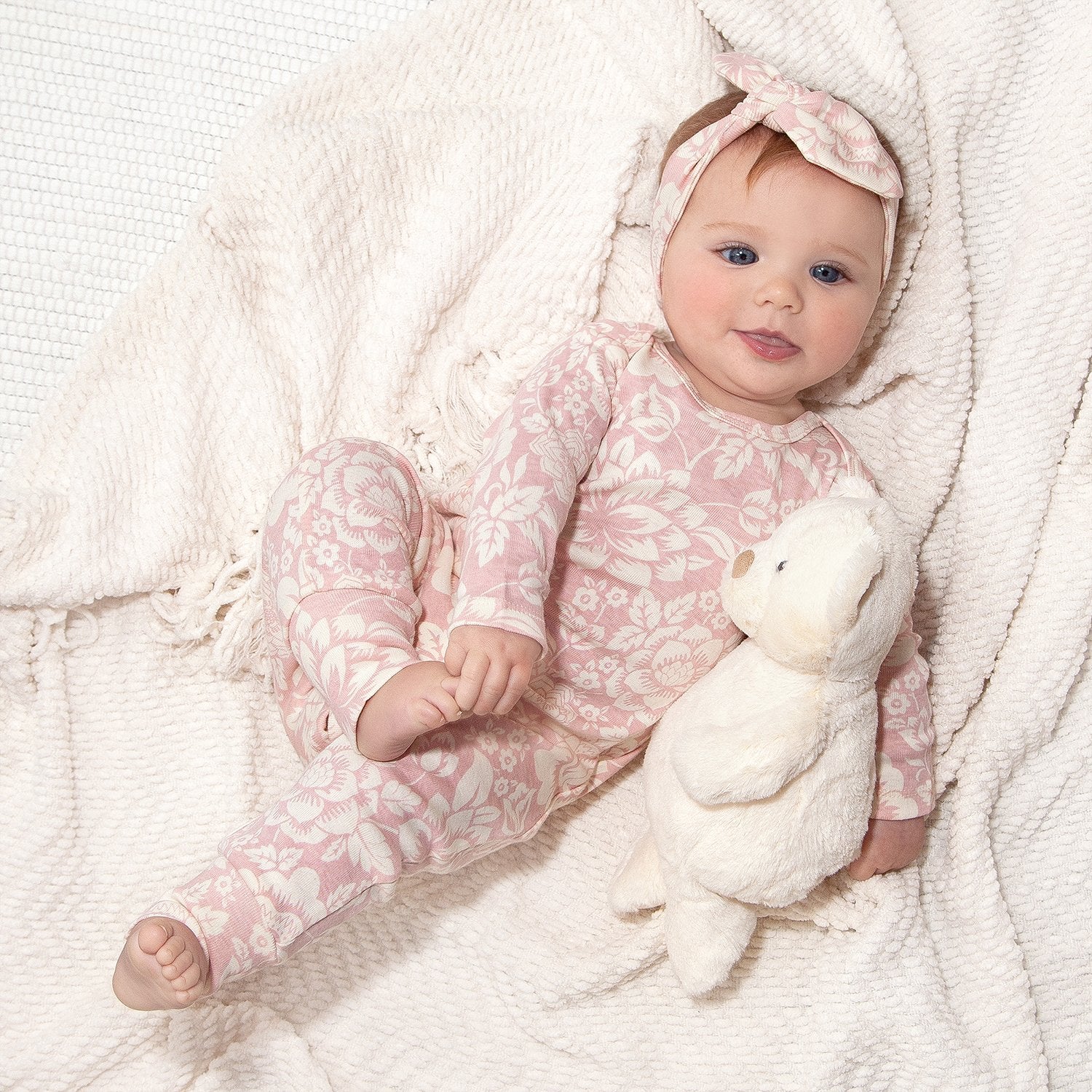Baby in pink floral outfit holding a white teddy bear on a soft white blanket