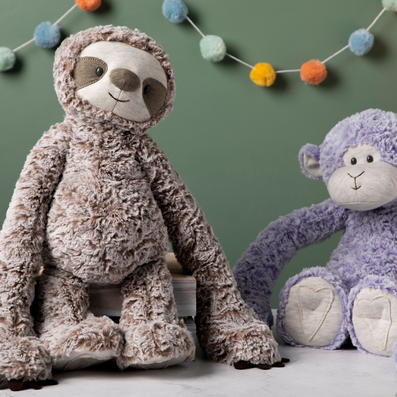 Two plush toys, one brown and one purple, sitting on a light surface with a string of colorful pom-poms in the background.